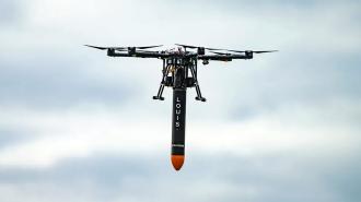 A drone in the air with a missile-shaped capsule hanging from its underside